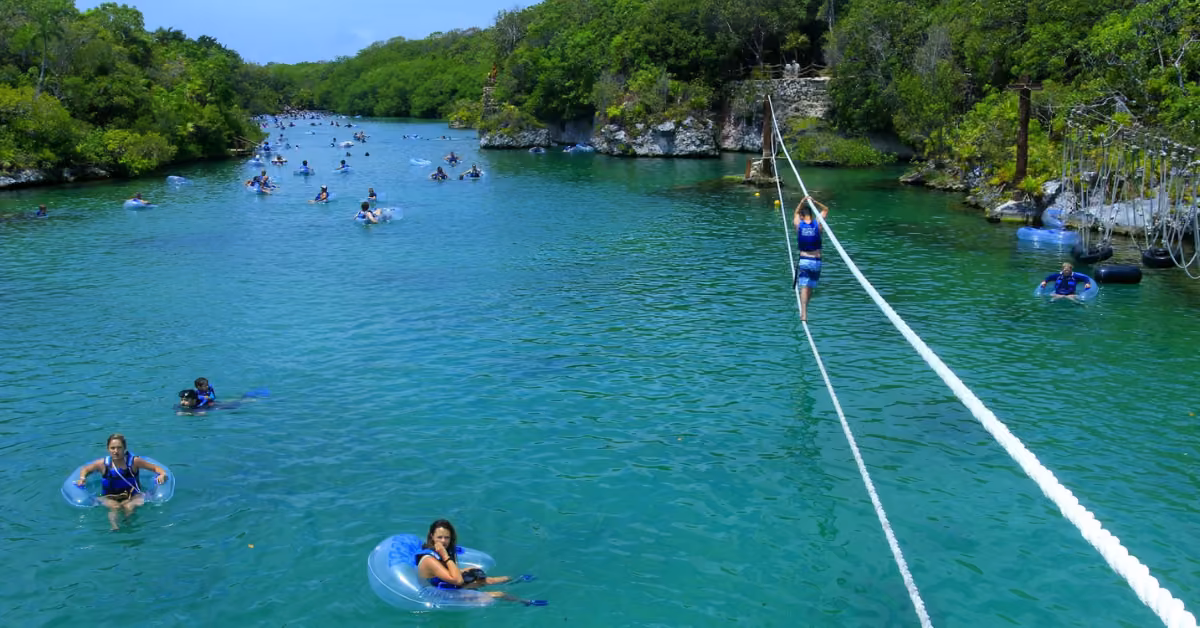 Visitors relax at Xel-Ha, floating on blue inner tubes or crossing clear river water surrounded by lush green scenery.