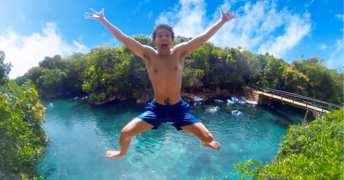 A man in blue swim trunks jumps into a clear, blue-green pool at Xel-Ha, backed by lush greenery and a wooden bridge.