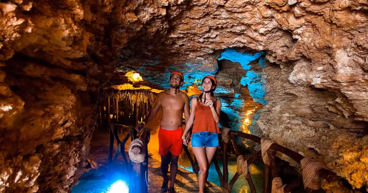 A man and woman in helmets walk through the glowing Xel-Ha cave, past rocky walls, wooden railings, lights, and stalactites.