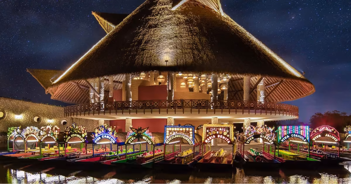 A large thatched-roof building glows at night as colorful boats sit docked on the water beneath a starry sky.