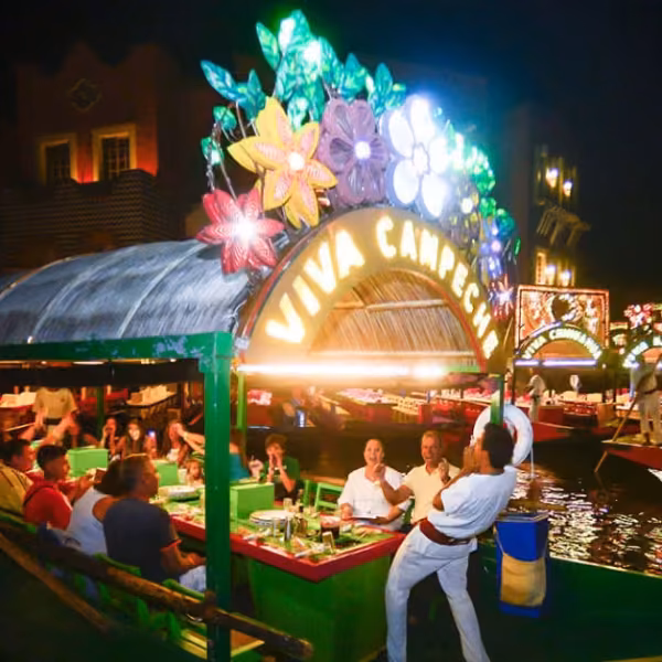 A lively boat with flowers and a Viva Chaparrita sign carries diners along a lit canal at night, with more boats behind.