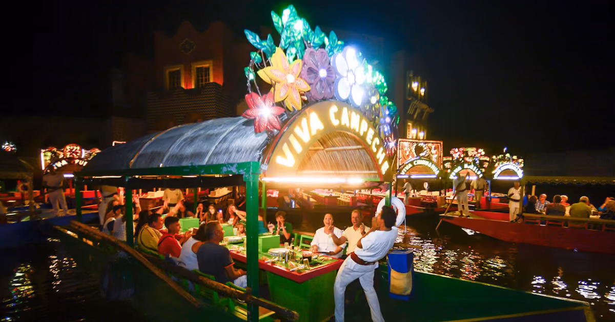 A lively boat with flowers and a Viva Chaparrita sign carries diners along a lit canal at night, with more boats behind.