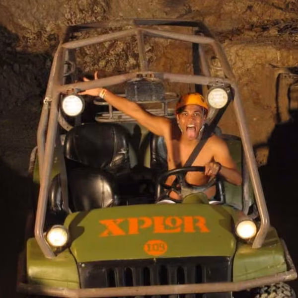 Helmeted driver steers an Xplor off-road vehicle through a rocky cave, arm raised and tongue out as headlights light the walls.