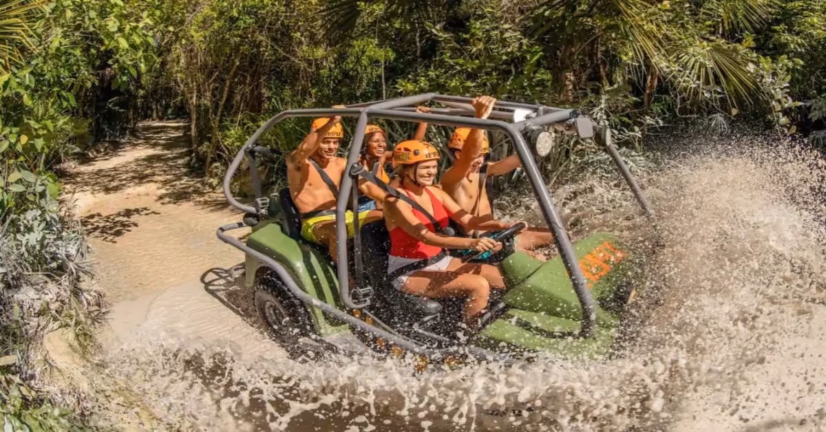 Four helmeted riders in bright clothes drive an open ATV through a muddy jungle trail, splashing water amid lush greenery.