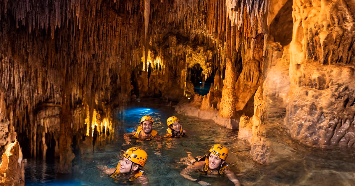 Four people in yellow helmets swim through a clear, narrow cave, rocky walls and stalactites lit by warm artificial light.