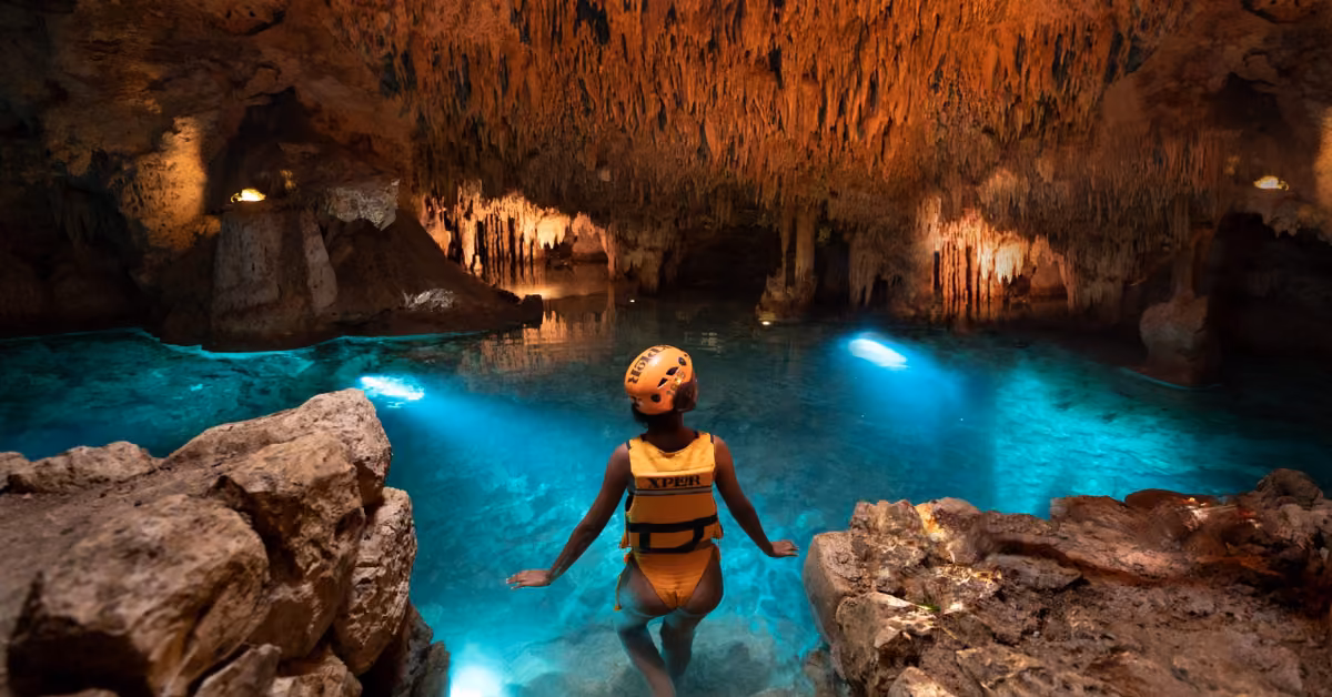 A person in a helmet and life jacket stands in clear blue cave water, surrounded by illuminated rock formations and stalactites.