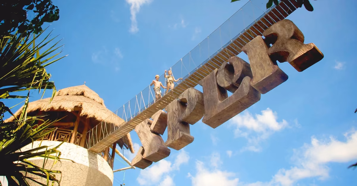 Two people sit on a hanging bridge above stone Xplor letters near a thatched tower, enjoying scenic views under a blue sky.