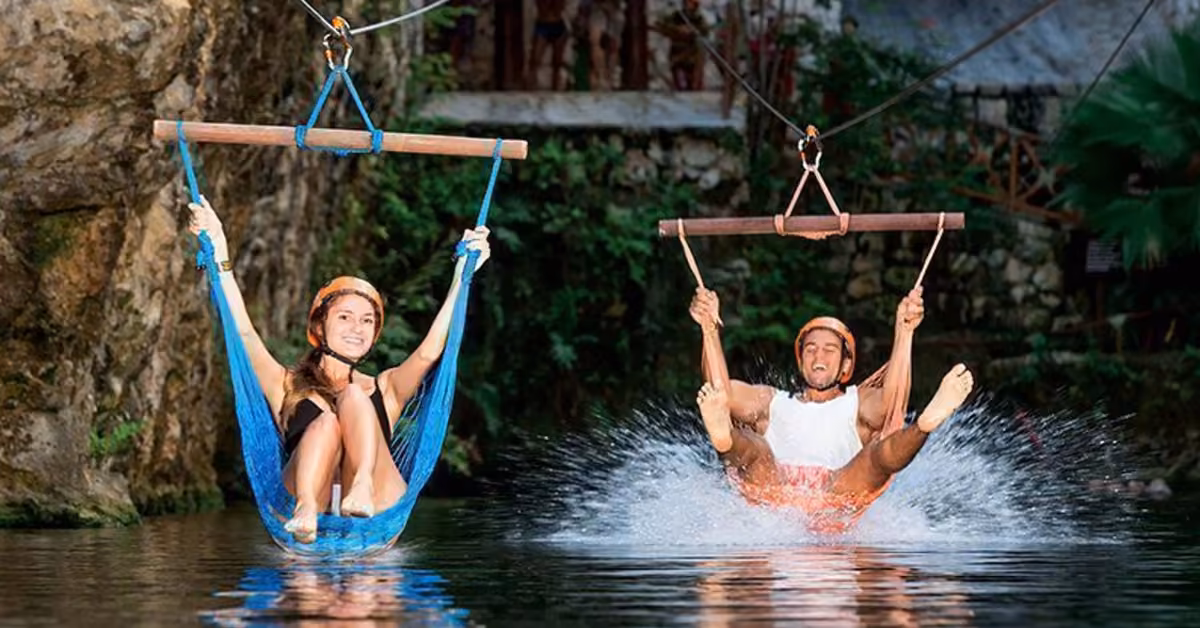Two people in helmets ride ziplines over water at Xplor, smiling and splashing, amid rocks and lush green scenery.