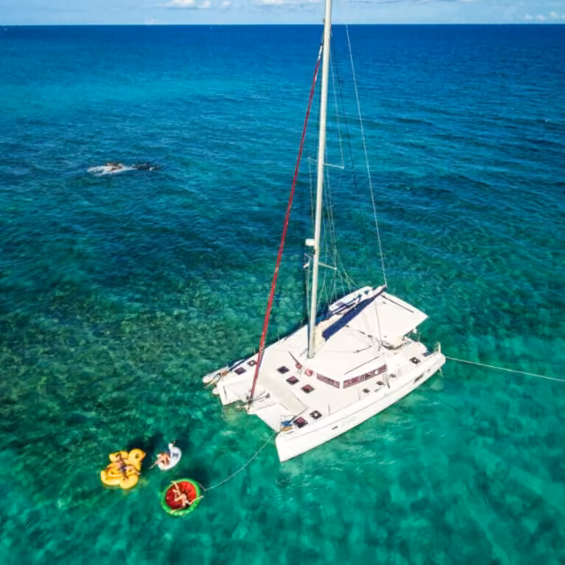 A white catamaran floats in clear turquoise water as people relax on inflatables; the ocean glimmers under a blue sky.