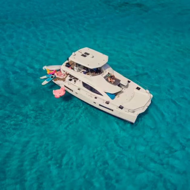A white yacht drifts on turquoise water, with pool floats nearby as people relax onboard and enjoy a sunny, carefree day.