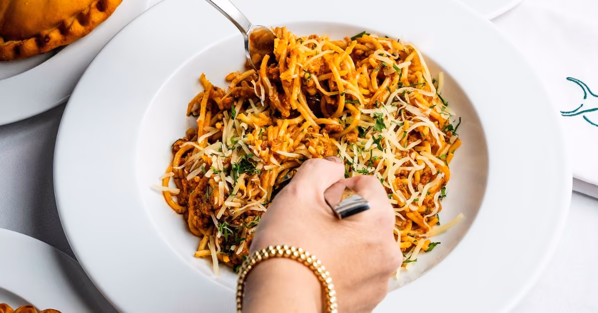 A hand with a gold bracelet twirls spaghetti topped with tomato sauce and cheese in a white bowl, garnished with herbs.