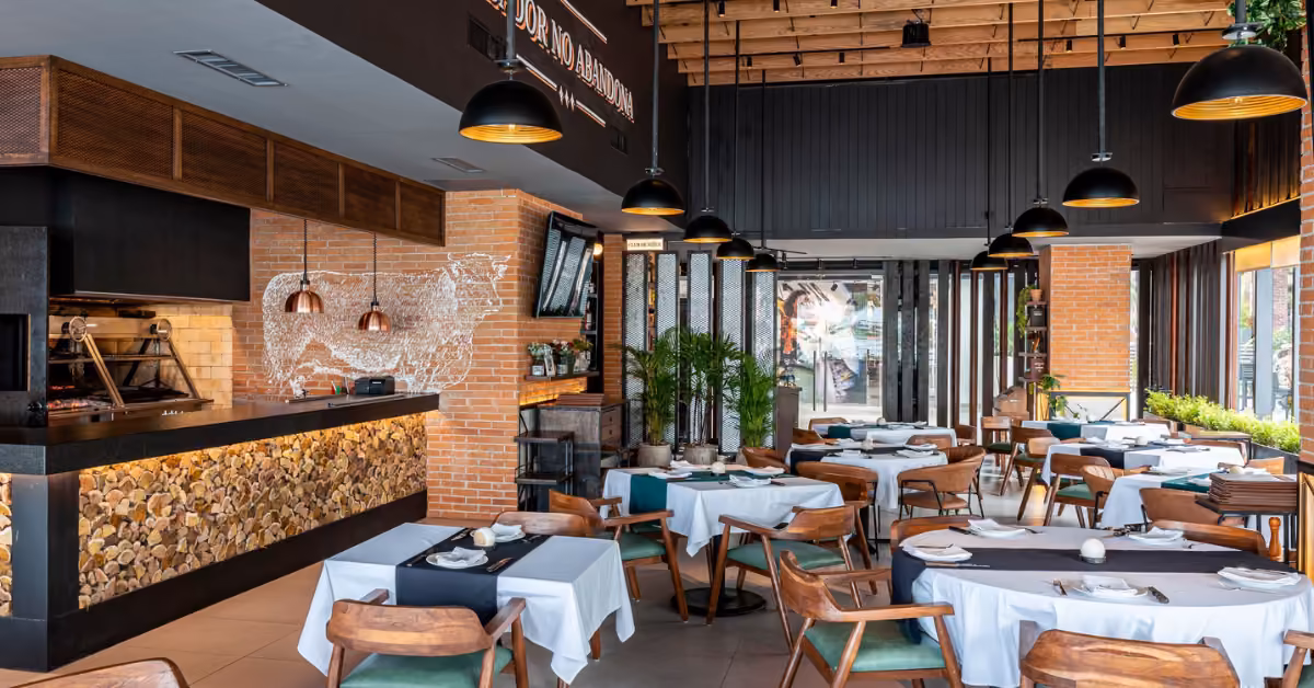 Cozy restaurant with wooden chairs, brick walls, white-black tablecloths, hanging lamps, stone counter, plants, and big windows.