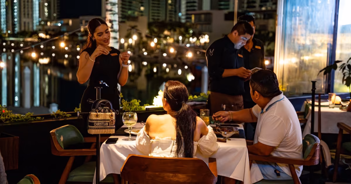 A woman chats with a couple at an outdoor restaurant table at night as a waiter prepares nearby, city lights glowing behind them.