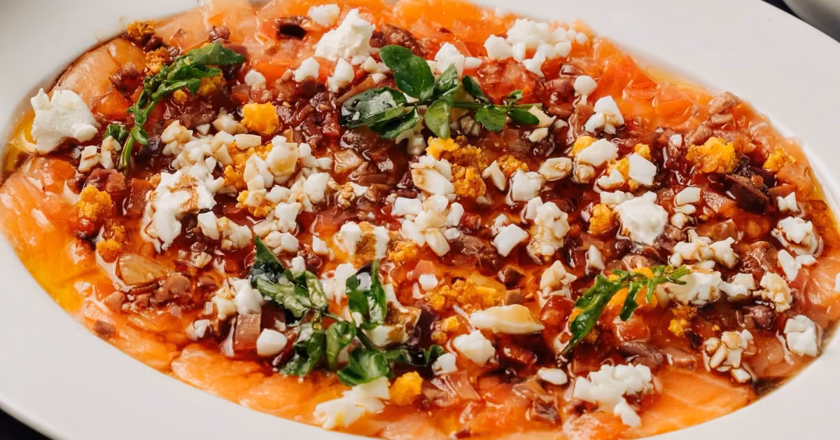 Thin salmon carpaccio on a white plate, topped with crumbled cheese, chopped nuts, greens, and bits of hard-boiled egg.