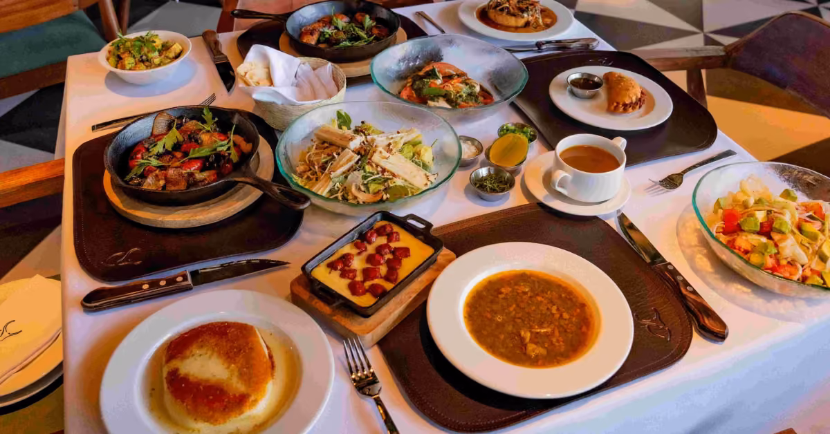 An elegant table displays salad, soup, grilled meats, bread, and desserts on plates and bowls over a white tablecloth.