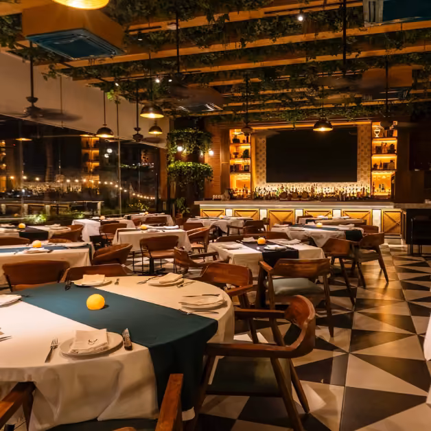 Warmly lit restaurant with wooden chairs, white tablecloths, green runners, checkered floor, hanging plants, and a lit bar.