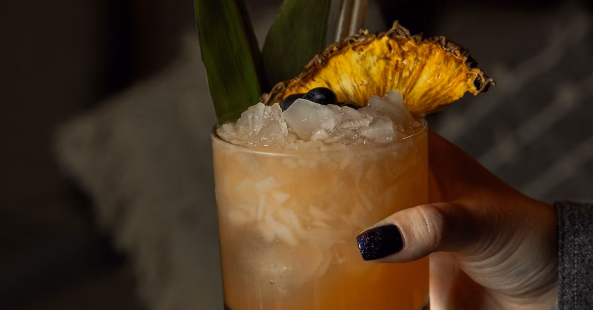 A hand with dark, glittery nails holds a cocktail garnished with crushed ice, dried pineapple, green leaves, and blueberries.