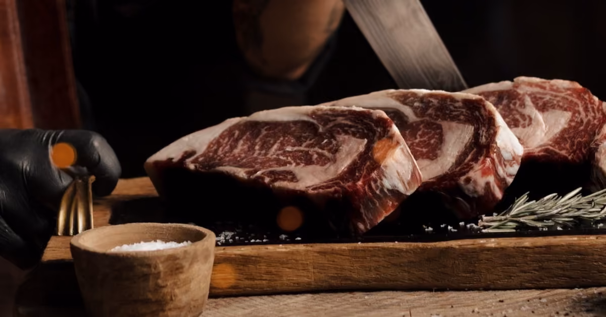 Three raw marbled steaks on a wooden board with rosemary and salt; a person holding a knife is in the background.