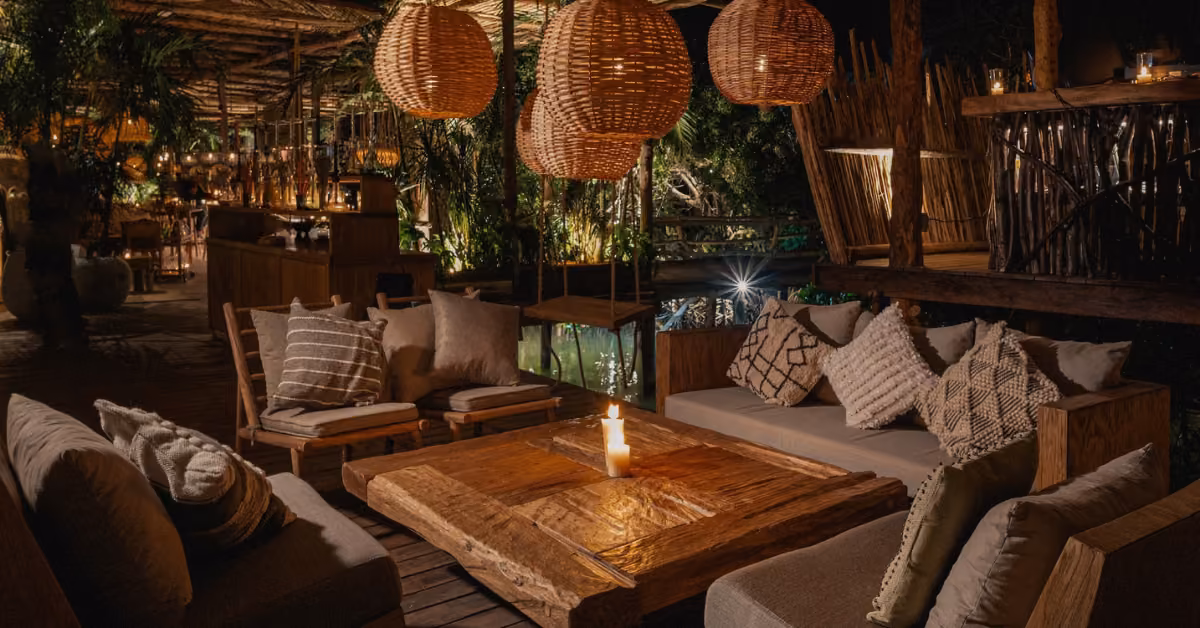 Cozy outdoor lounge at night with wooden furniture, cushioned sofas, pendant lights, pillows, and lush greenery.