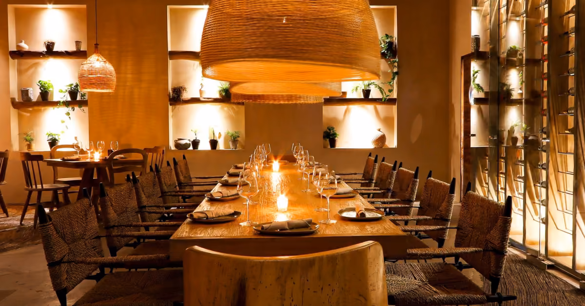 A cozy restaurant dining room with a long wooden table, woven chairs, wicker lamps, candles, and shelves with plants and pottery.