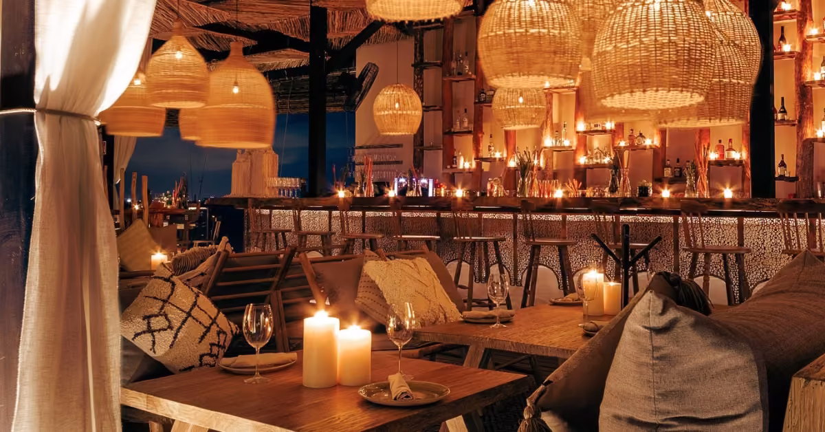 Chambao restaurant with wooden tables, cushioned chairs, wicker lamps, candlelit tables, and a decorated bar in the background.