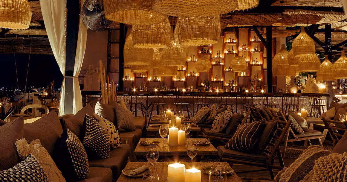 A cozy, dim restaurant with woven lamps, candlelit wooden tables, plush sofas, and shelves glowing warmly at night.