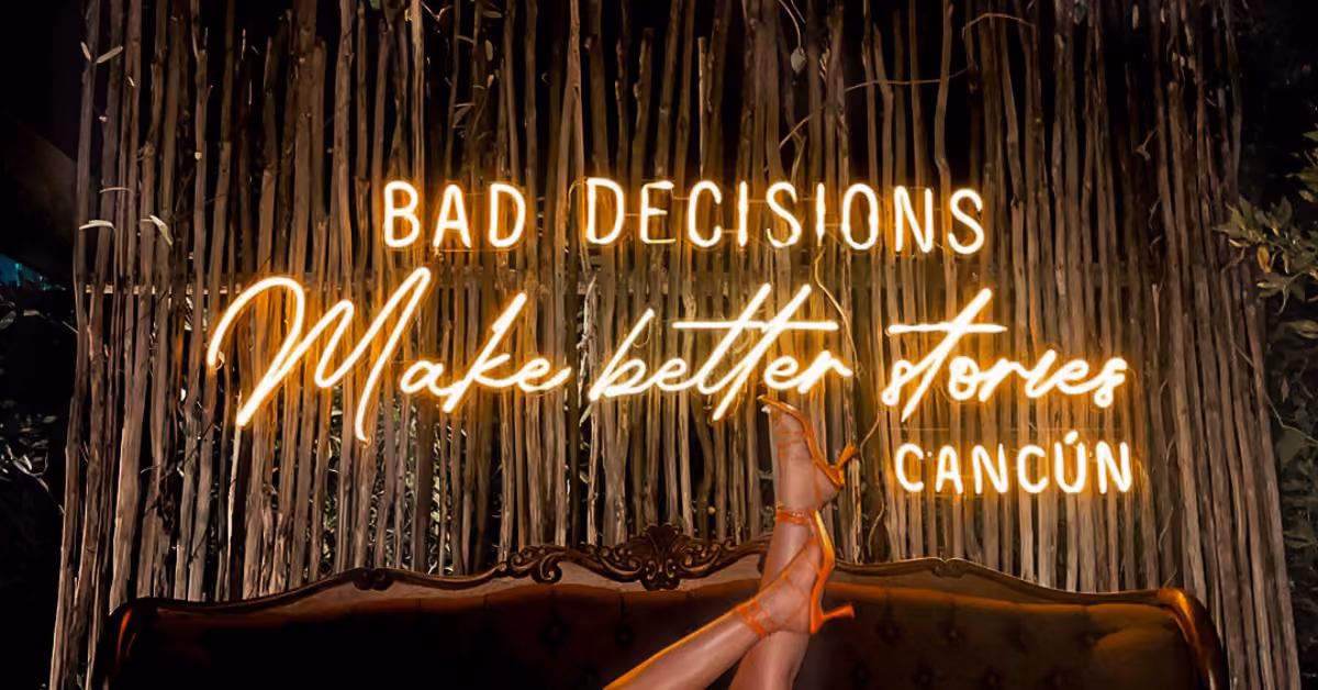 Neon sign says "Bad decisions make better stories" above a vintage sofa, with legs in heels playfully up and bamboo backdrop.