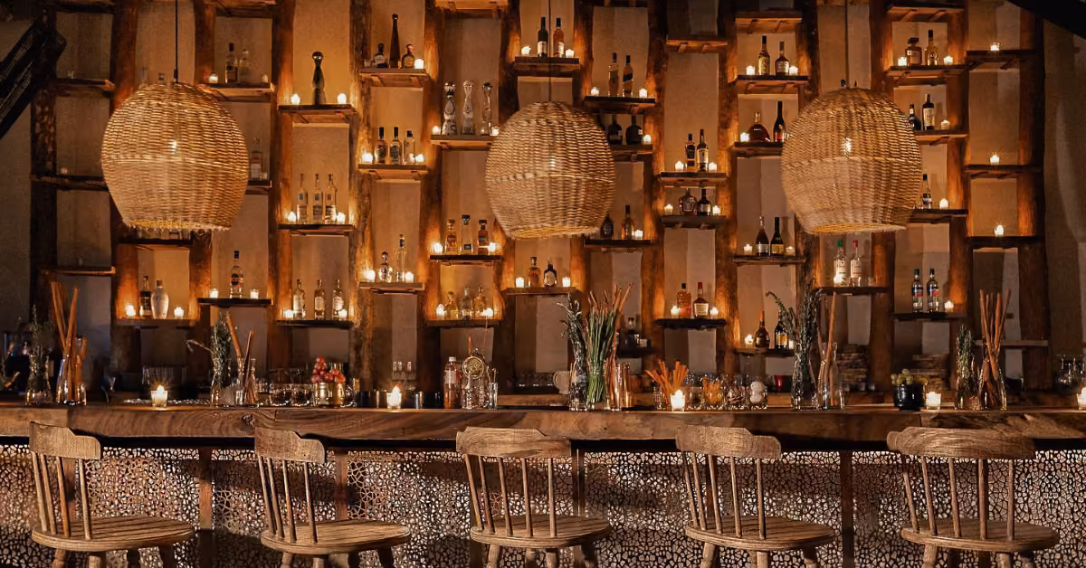 A cozy bar with wooden stools, a log-accented counter, bottles on shelves, and soft lighting from candles and wicker pendants.