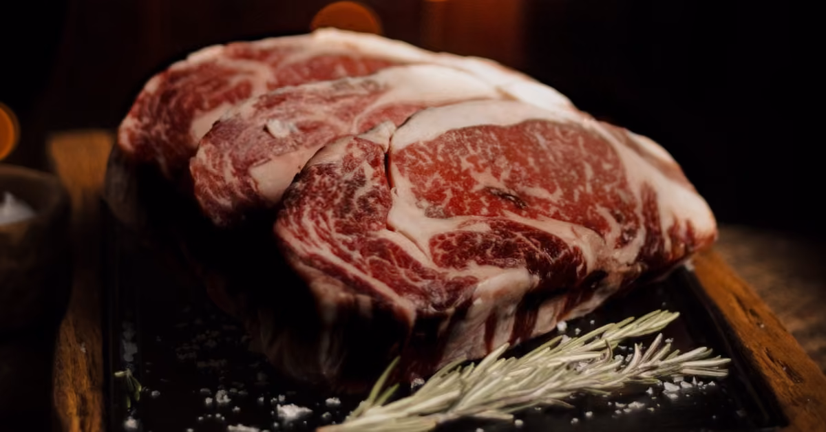 Two marbled raw steaks on a dark tray, topped with coarse salt and rosemary, their rich colors highlighted by a blurred background.