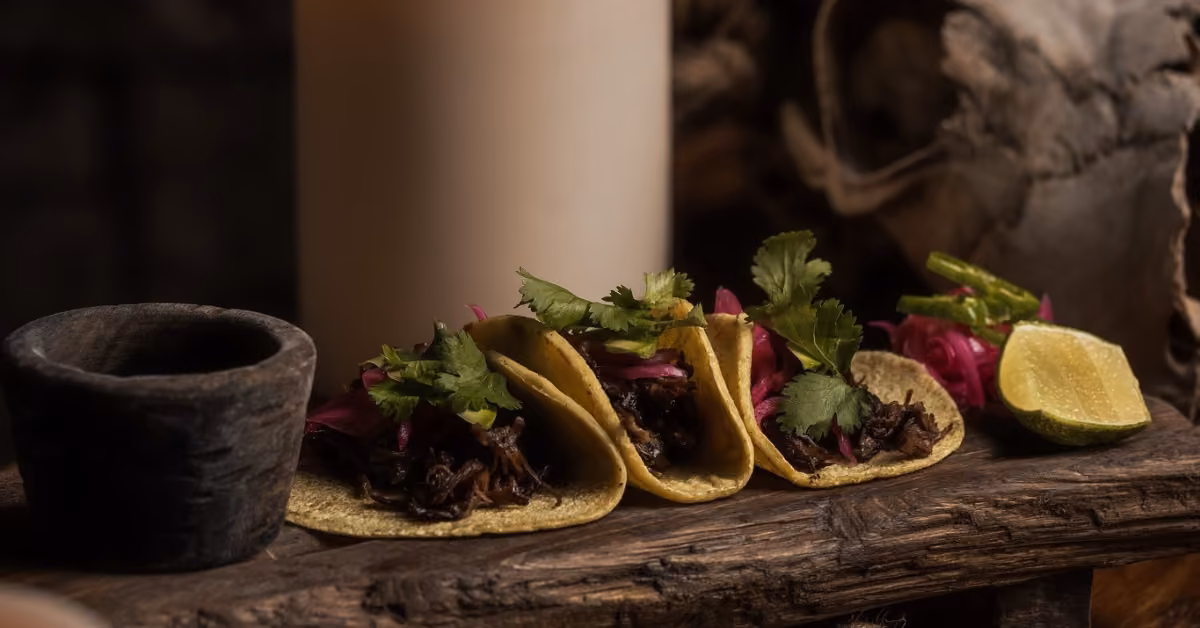 Three tacos with shredded meat, cilantro, and red onions on a wooden table, served with lime and a small stone bowl.