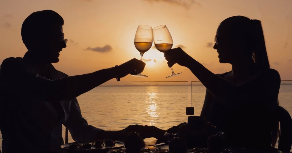 A couple sits by the water at sunset, silhouetted as they toast and hold hands across a table with food and drinks.