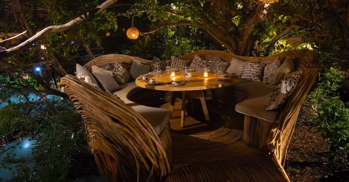 Round wooden table with candles, cozy woven benches, and glowing lanterns under trees creates a warm outdoor setting.