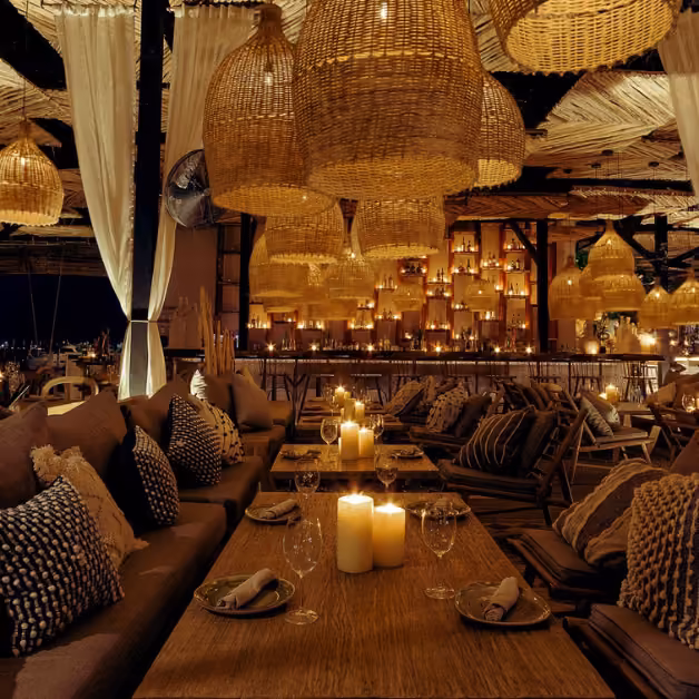Cozy restaurant interior with wooden tables, cushioned chairs, candles, rattan lamps, soft golden light, and white draped fabric.