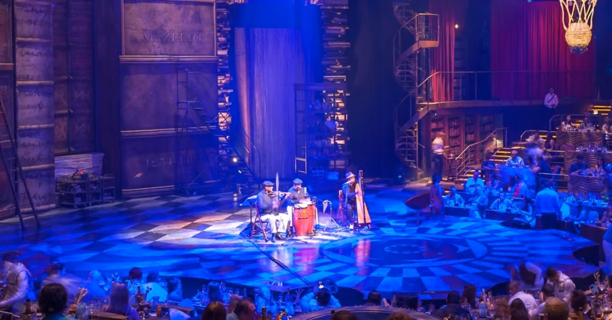 Musicians perform on a vibrant circular stage with dramatic lights, spiral stairs, and bookshelves, as the audience sits at tables.