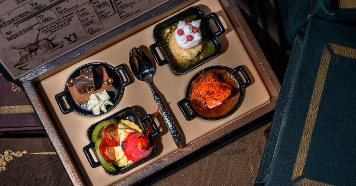 A tray with four small black dishes of gourmet desserts and a spoon sits atop old books and vintage paper with printed text.
