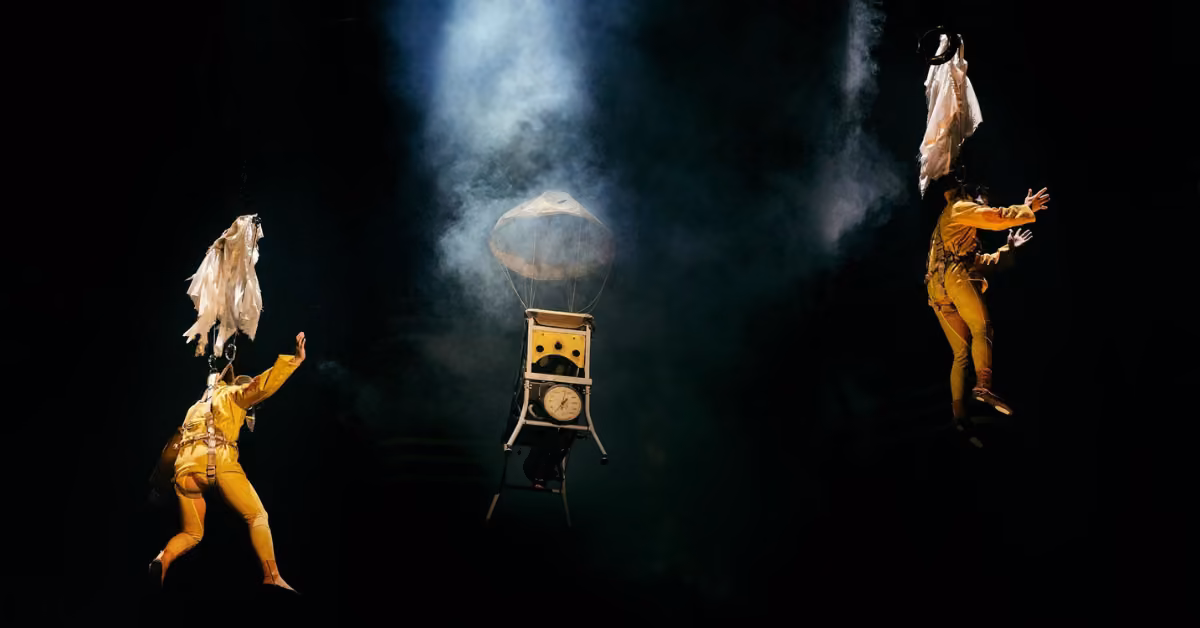 Two performers in yellow float mid-air on a smoky dark stage, with a vintage clock and lamp between them in a dreamlike scene.