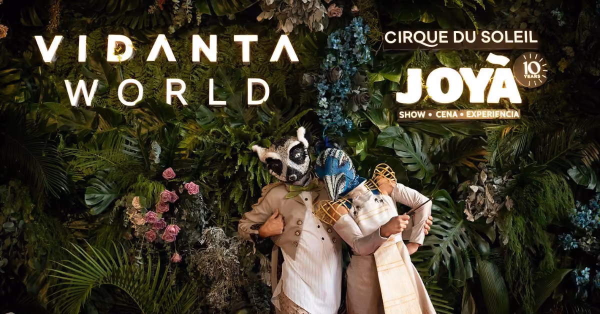 Two performers in animal masks and elegant costumes pose by a lush foliage wall with Cirque du Soleil logos for Joya’s 10th anniversary.