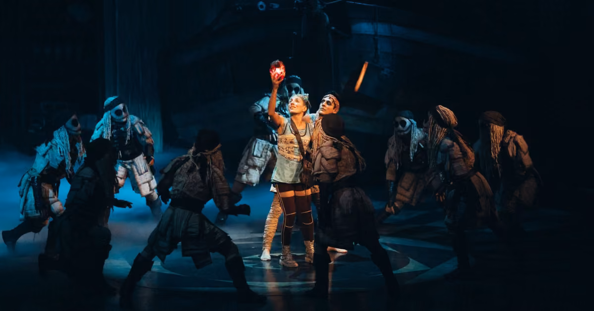On a dim stage, costumed performers gesture dramatically as one center figure lifts a glowing red object. A ship looms behind.