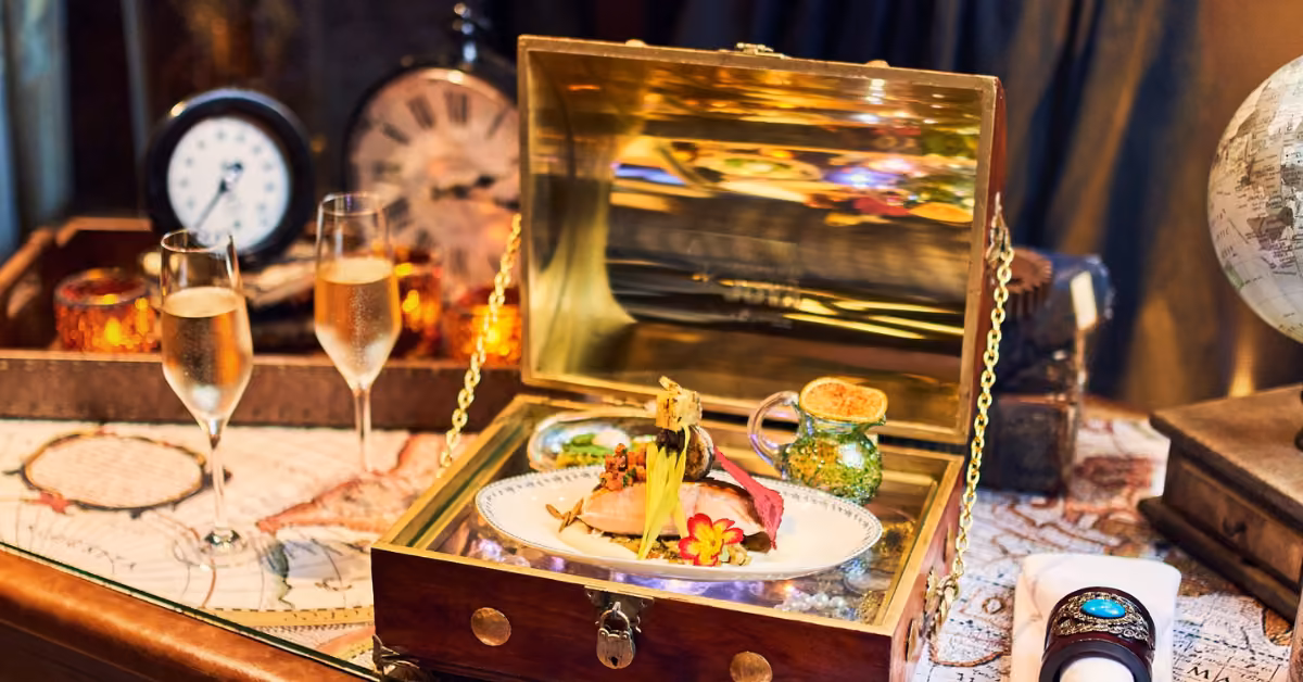 A decorative plate with a gourmet dish sits in an open chest, surrounded by champagne glasses, candles, clocks, and a map.