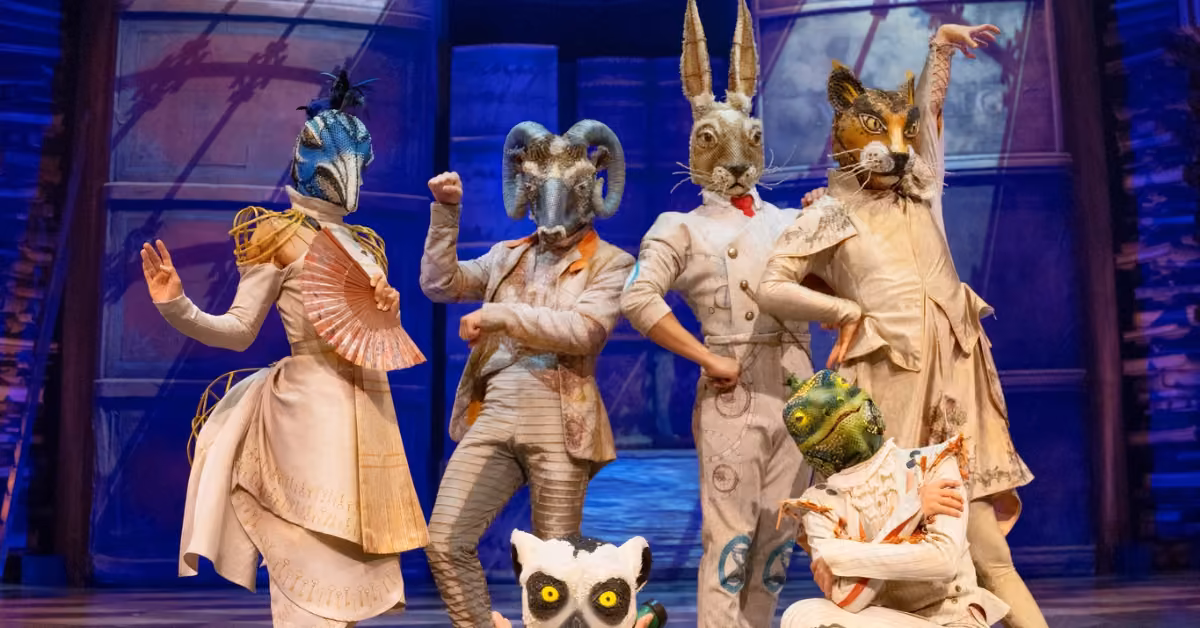 Performers in vibrant costumes and animal masks pose onstage, with a lemur-masked figure crouched in front amid vivid scenery.