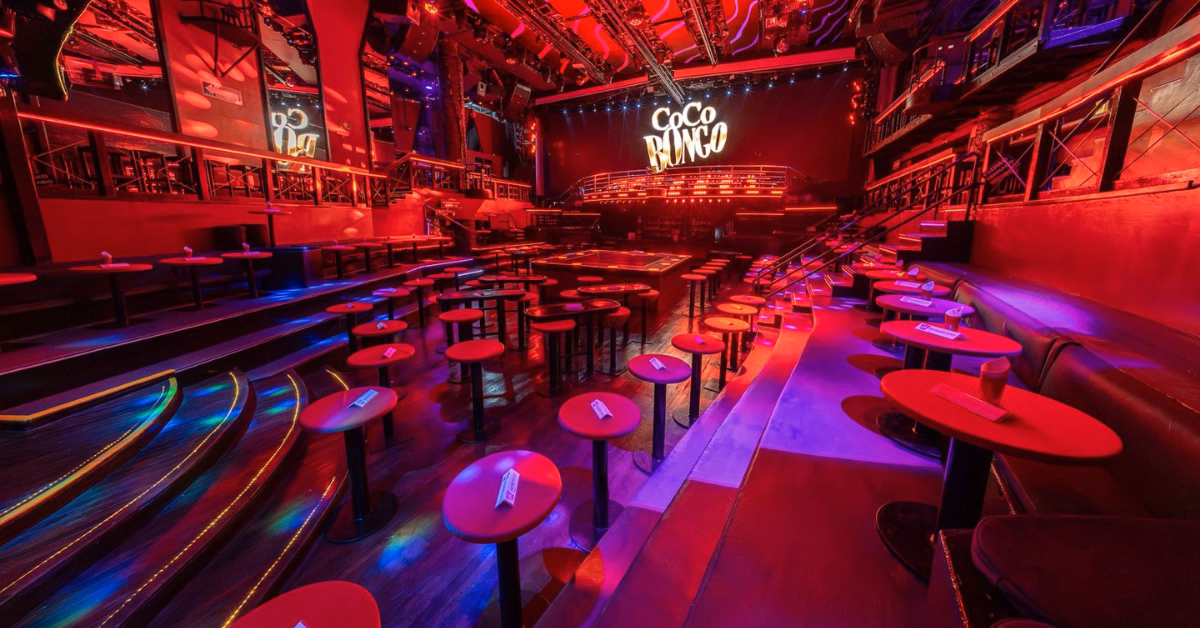 Coco Bongo Night Club with red lighting, round tables, and benches surrounds a lit stage featuring the Coco Bongo sign.