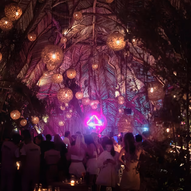 People dance indoors beneath glowing lanterns and a palm ceiling, bathed in soft pink and purple lights at a festive party.