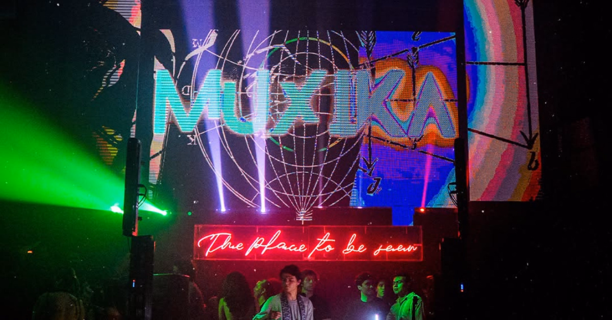 People gather in a lively club with colorful lights, a neon sign, and a screen displaying MUSIKA with artistic visuals.