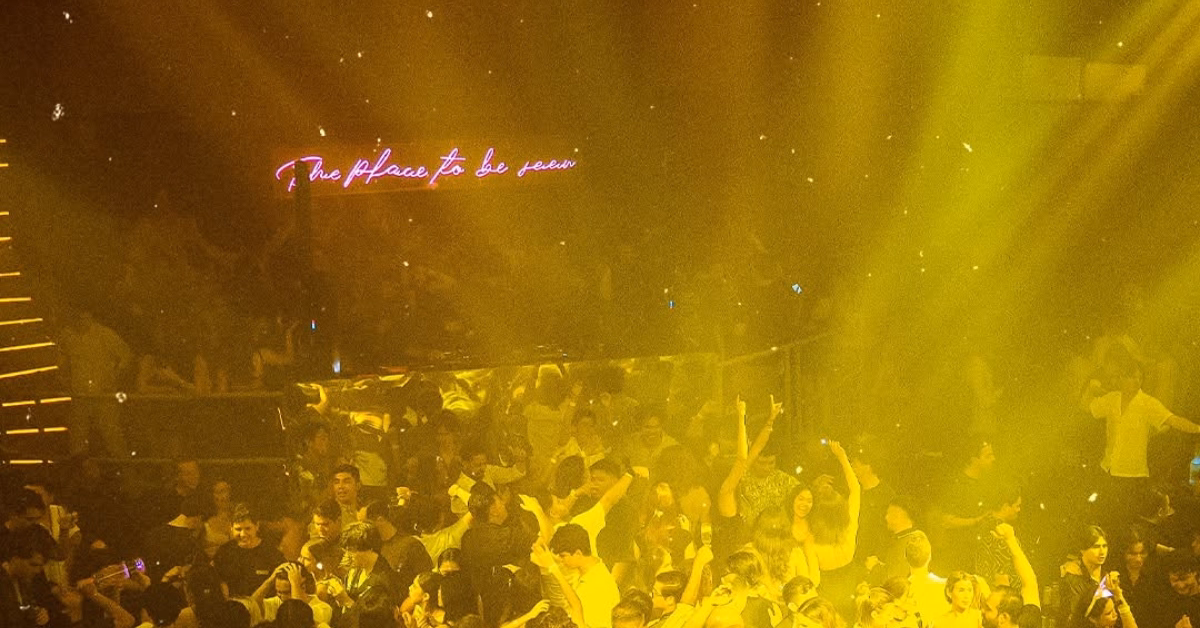 A lively nightclub with people dancing beneath yellow lights and a neon sign in the background reading “The place to be seen.”.