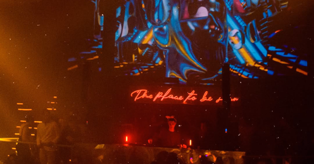 A dim nightclub with colorful lights, a DJ booth, neon sign, and people dancing by a vibrant screen above the DJ.