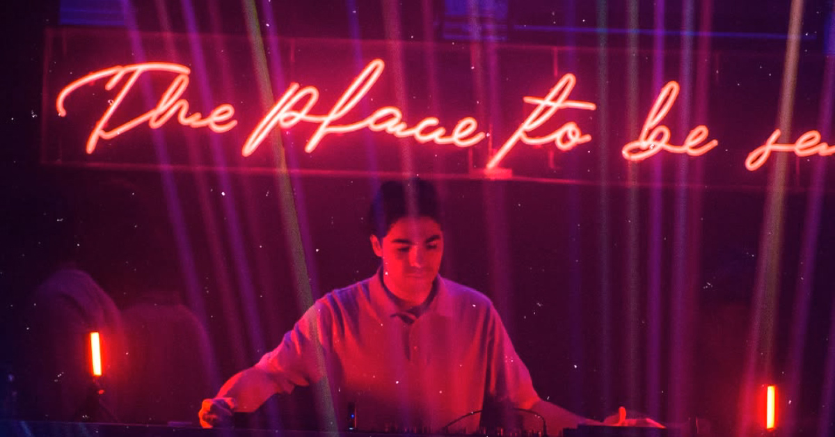 A DJ plays under pink neon lights reading “The place to be seen,” with purple rays creating a vibrant, club-like atmosphere.