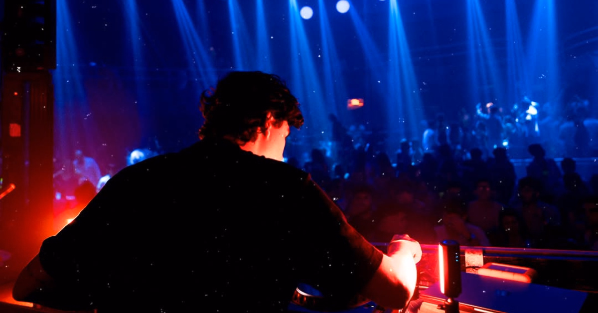 A DJ plays for a lively crowd in a dark nightclub, lit by red and blue lights as people dance in the background.