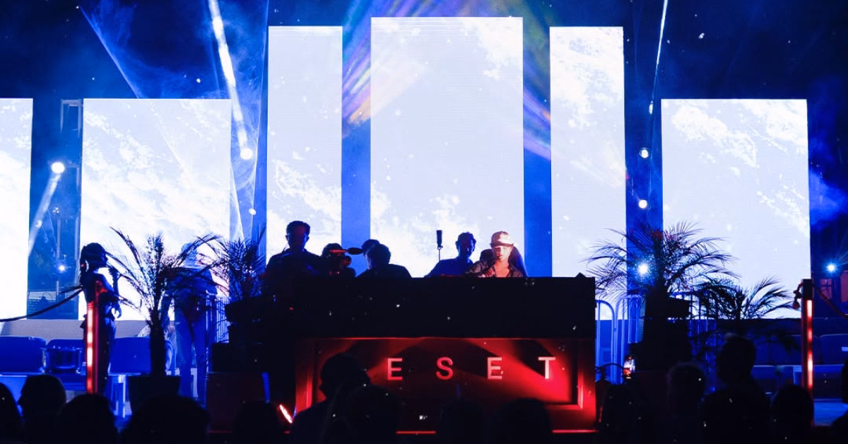 A DJ plays at d’cave nightclub, vibrant lights and palm plants around, as the word RESET glows red on the booth.