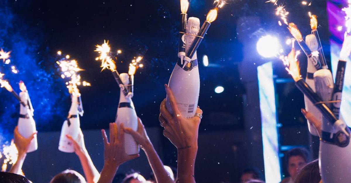 People lift champagne bottles with sparklers at a vibrant party under bright lights in a colorful nightclub.