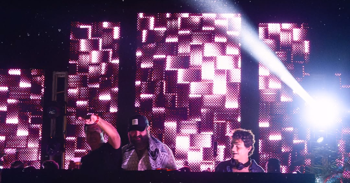 Two DJs perform at night at d’cave nightclub, lit by pink and purple LED lights. One points to the crowd as light beams behind.
